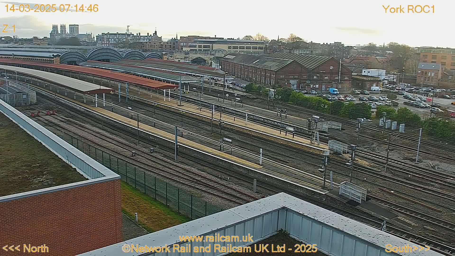 A high-angle, long shot of a train station on a partly cloudy day, showing numerous train tracks, platforms, and a large parking lot with cars.