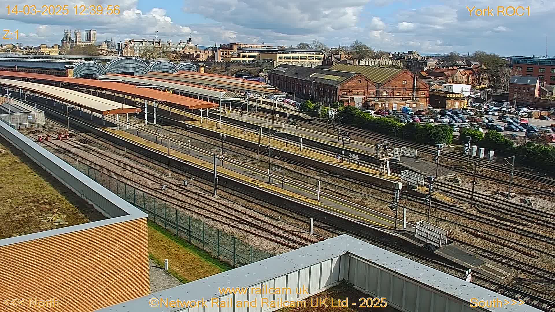 A high-angle, sunny view of a railway station with multiple tracks, platforms, and a large parking lot adjacent to brick buildings.