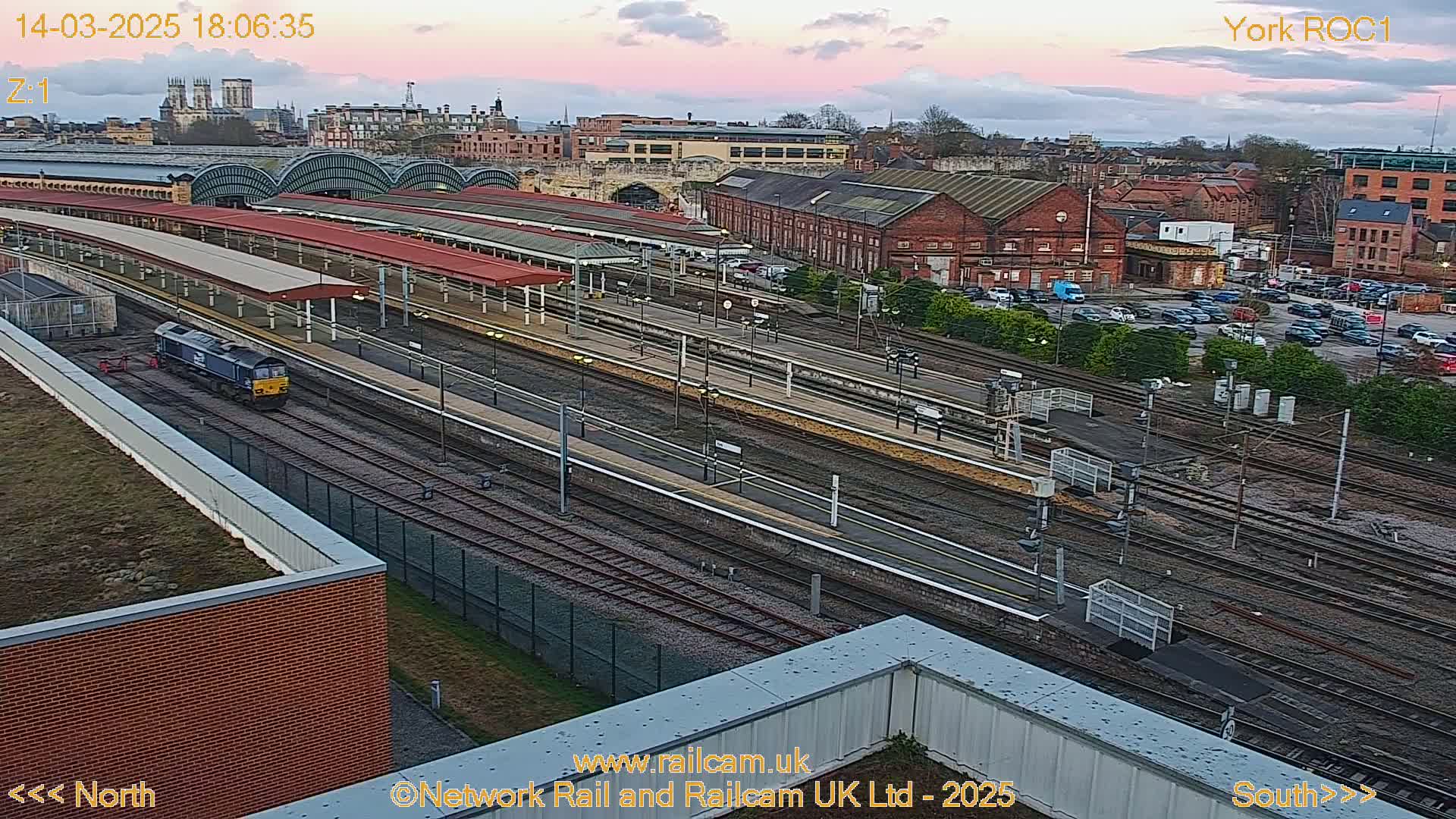 A train sits in a railway yard at dusk, under a partly cloudy sky, near a large train station and city buildings.