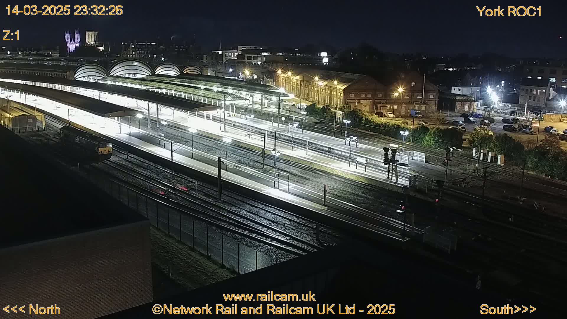 A nighttime, high-angle view of a brightly lit train station with multiple tracks and platforms, a locomotive visible at one end, and surrounding buildings and parking areas.