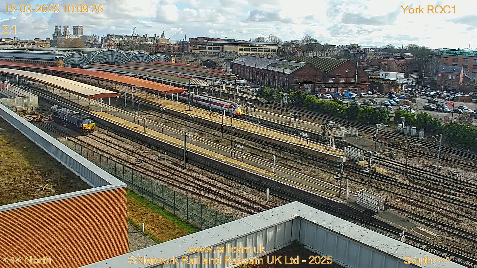 A high-angle view of a train station on a partly cloudy day, showing multiple train tracks, platforms with trains, and a city in the background.