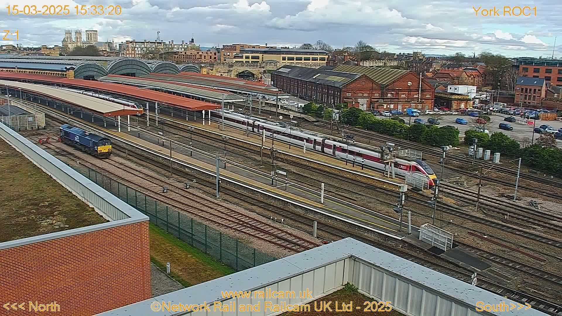 A high-angle view of a train station on a partly cloudy day, showing multiple trains on various tracks, with a city skyline visible in the background.
