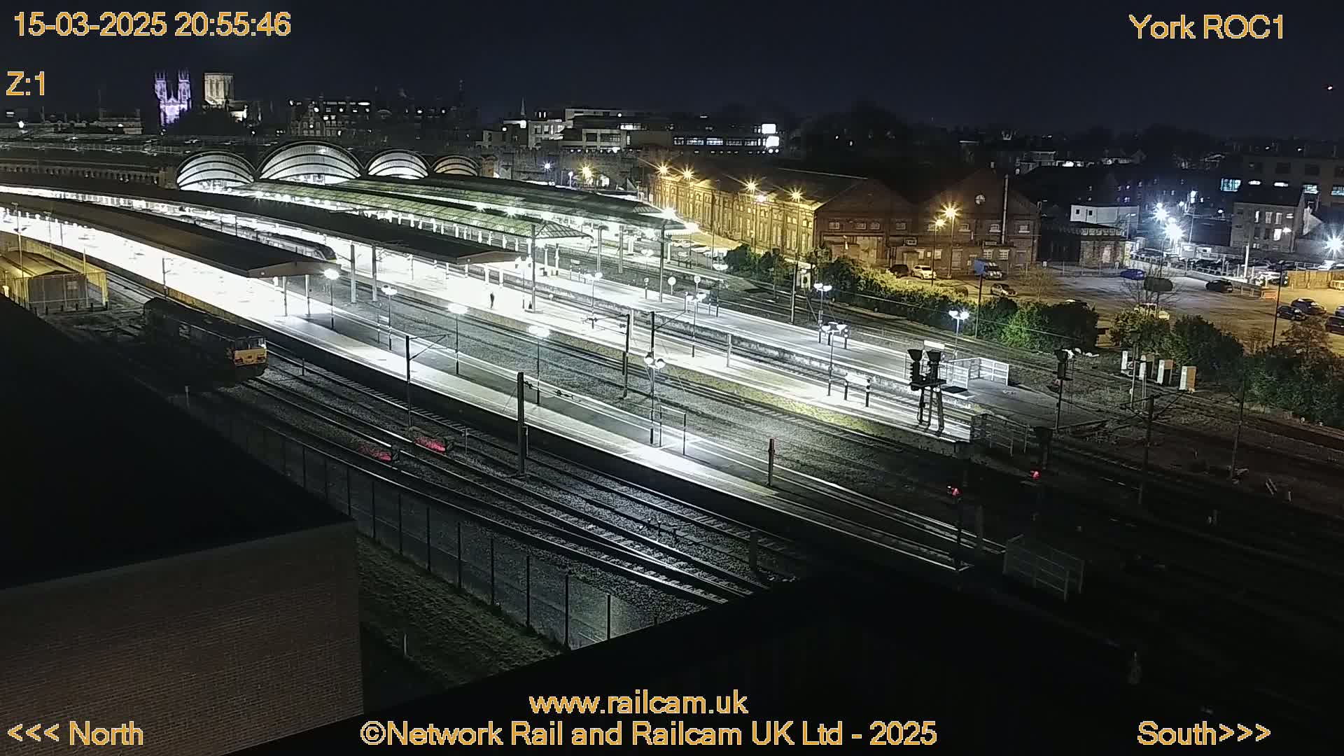 A nighttime, high-angle view of a train station with multiple tracks, platforms, and a train at the platform.