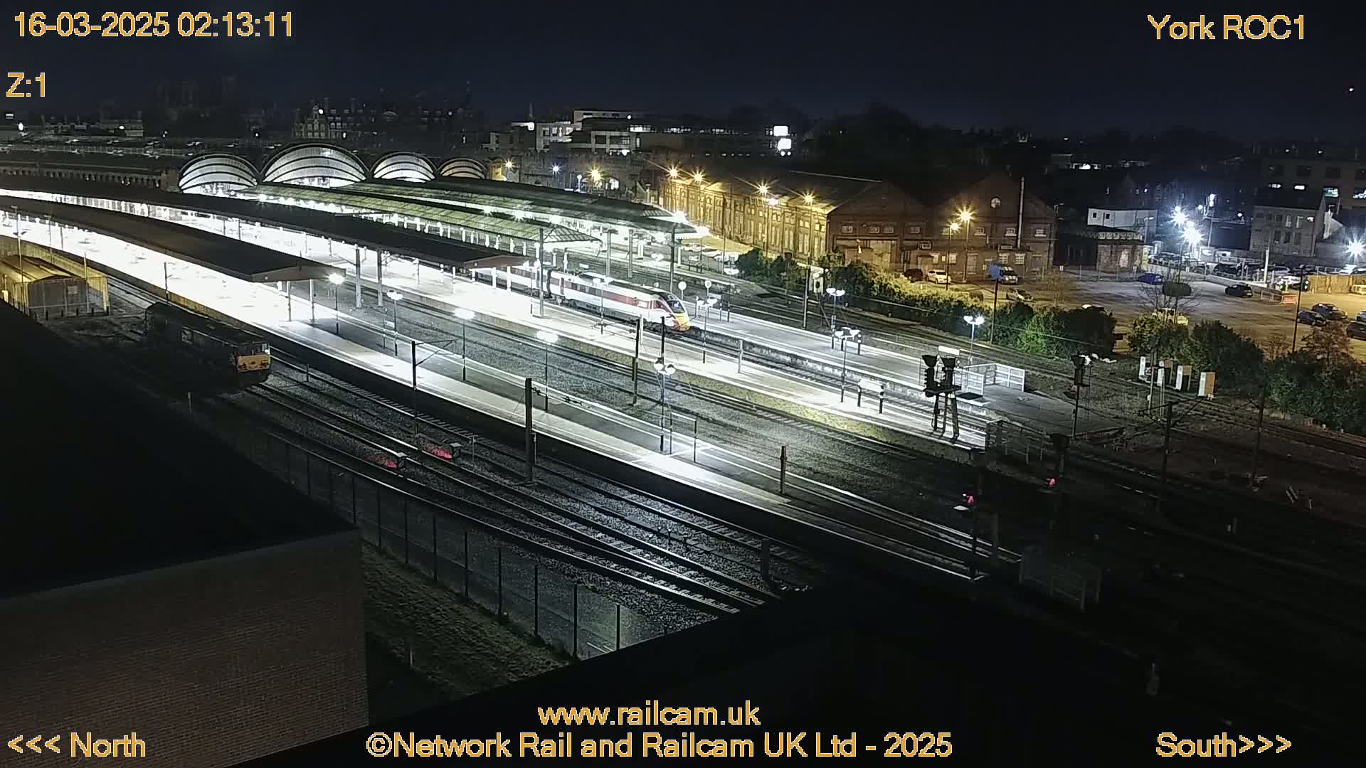 A nighttime, high-angle view of a train station with two trains on the tracks and illuminated platforms.