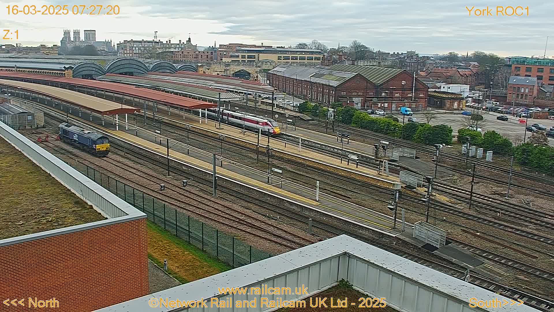A high-angle, long shot of a train station on an overcast day, showing multiple train tracks, platforms with red roofs, and several trains, including a diesel locomotive and a high-speed train.