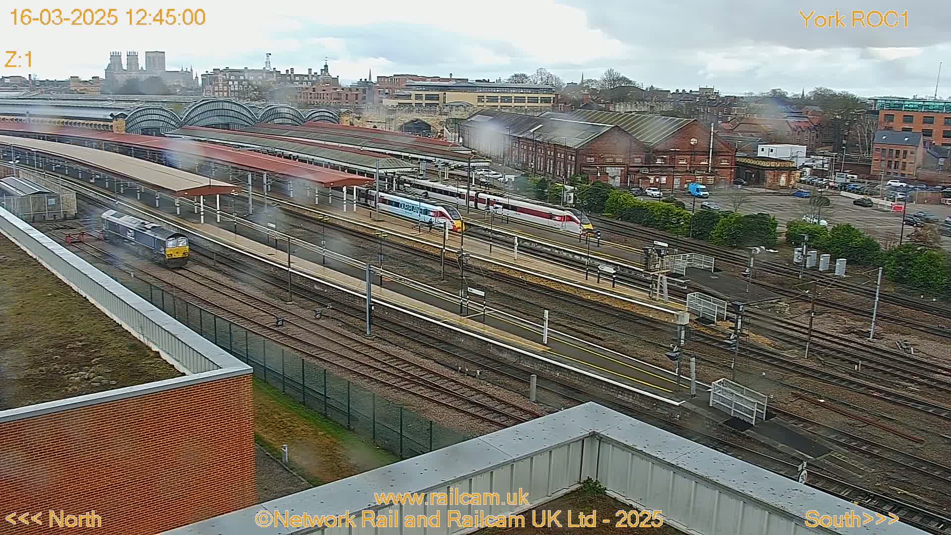 A high-angle view of a train station on an overcast day shows several trains on the tracks and a large station building in the background.