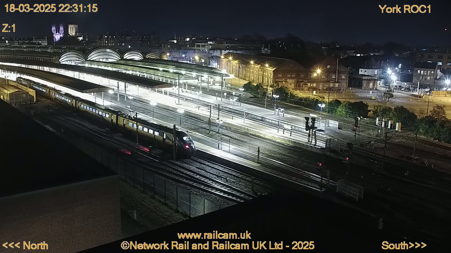A nighttime high-angle view of a train station with two trains on the tracks, illuminated by artificial lights.