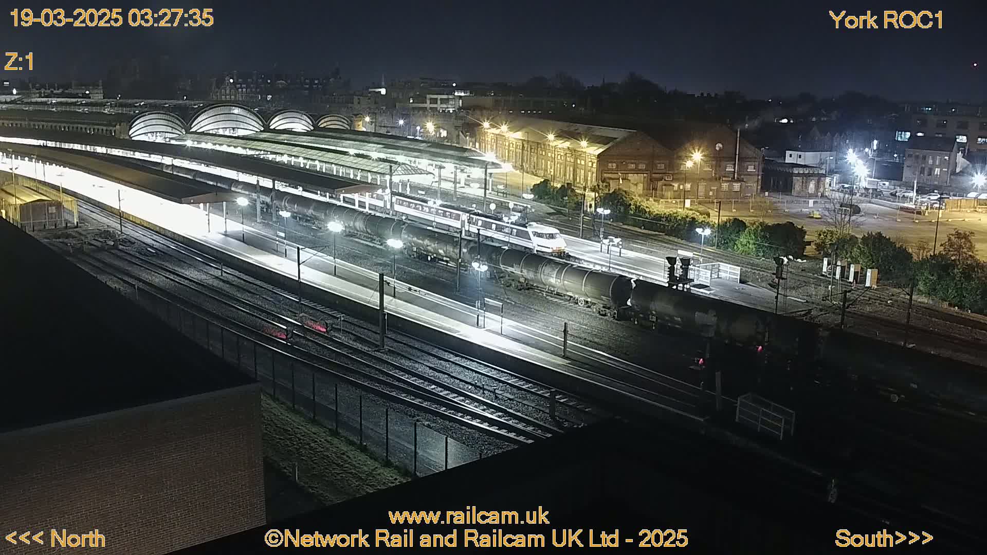 A nighttime, high-angle view of a train station with a passenger train and a freight train, illuminated by artificial lights.