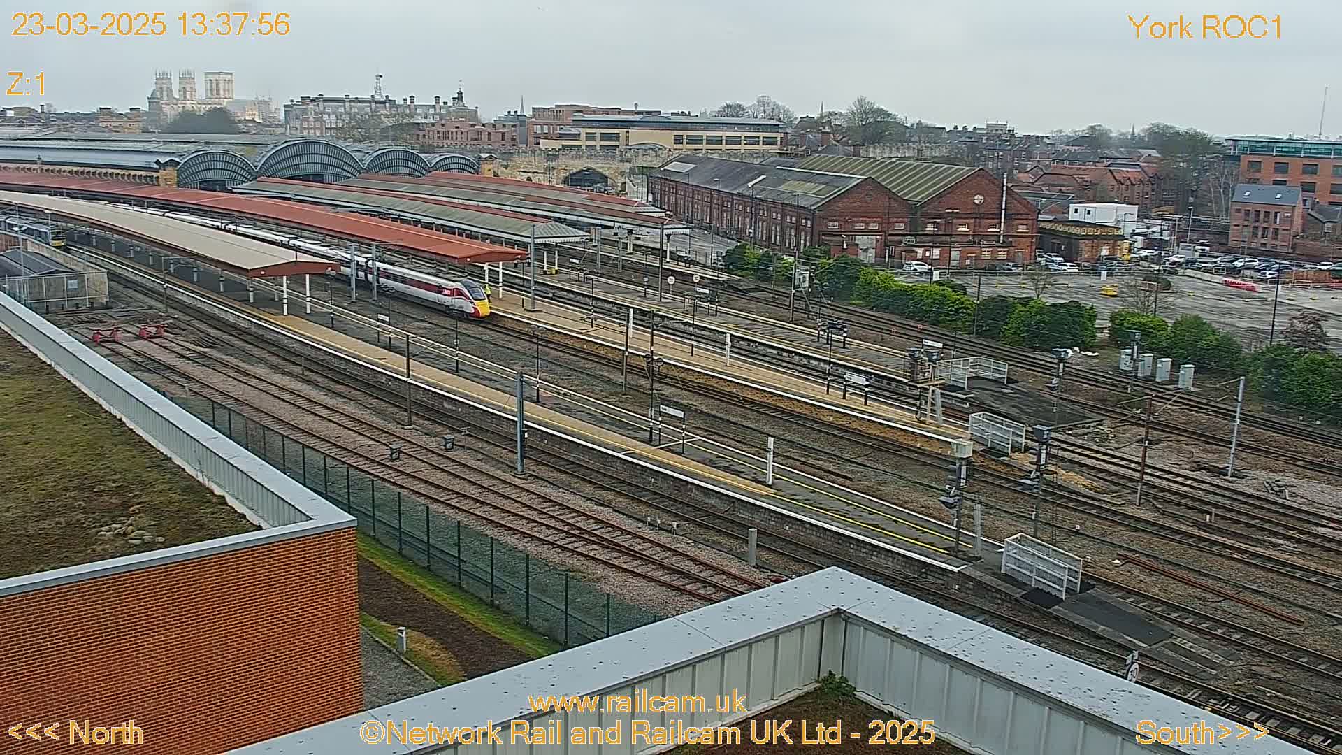 A high-angle view of a train station on an overcast day, showing multiple train tracks, platforms, and a high-speed train passing through.