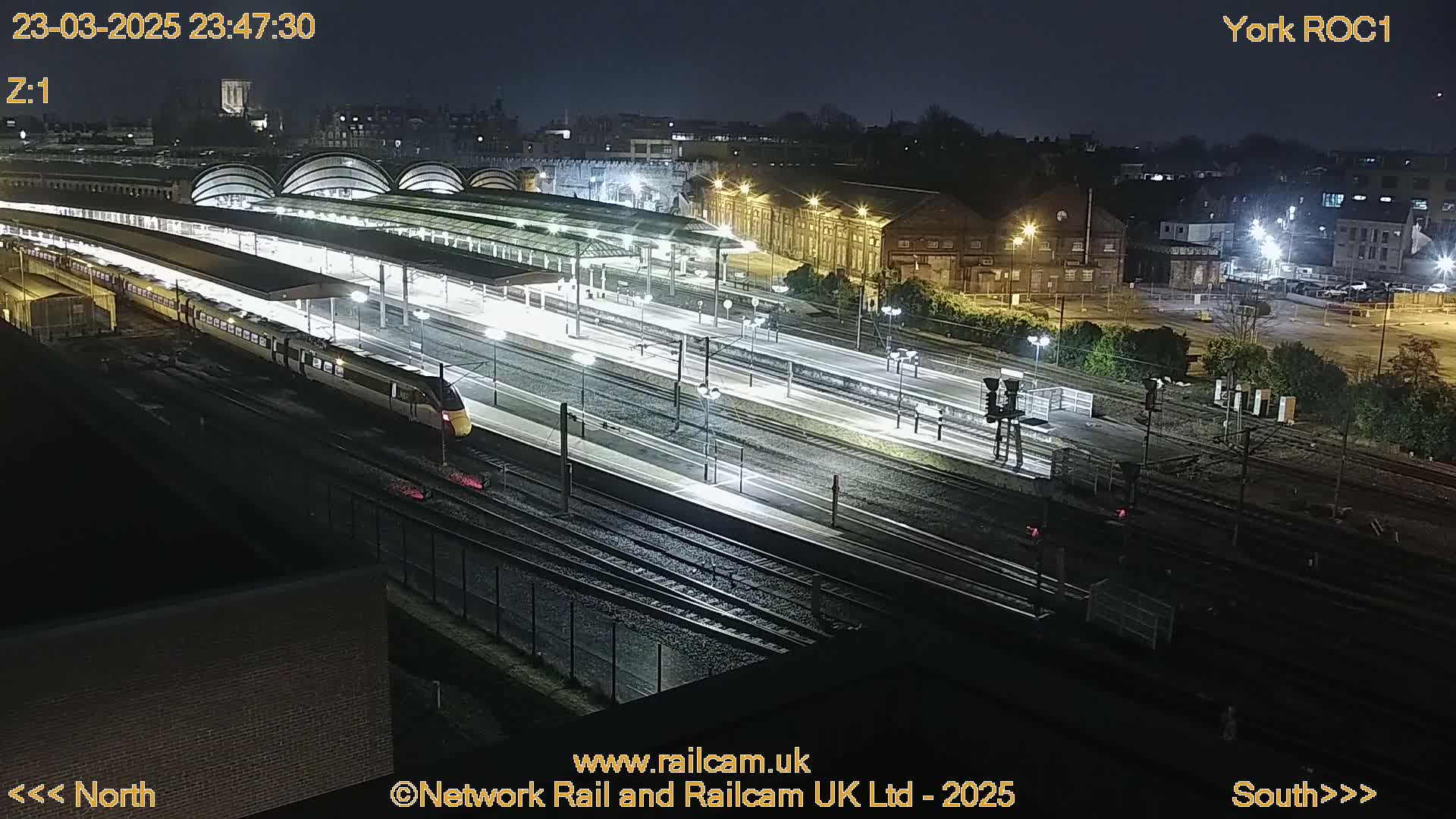 A high-angle nighttime view of a train station with a high-speed train departing, illuminated by artificial lights.