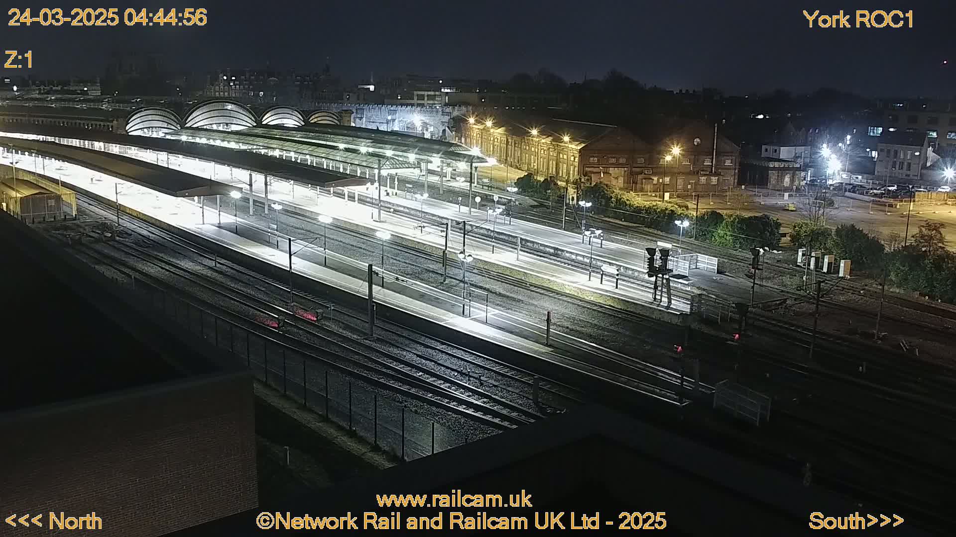 A nighttime, overhead view of a train station with numerous tracks and platforms illuminated under artificial lights.