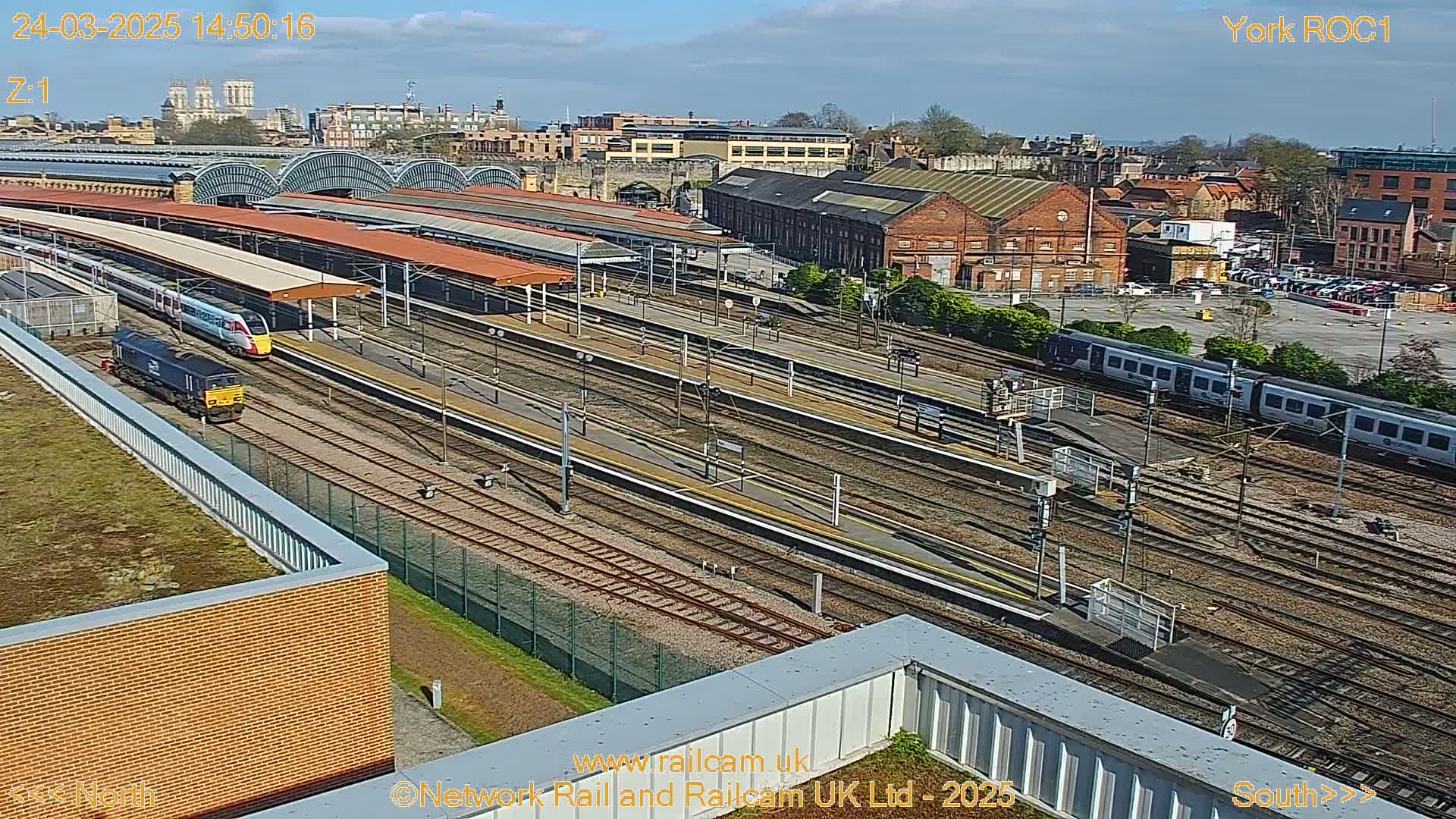 A high-angle, sunny view of a train station with multiple train tracks, platforms, and several trains, some of which are in motion.