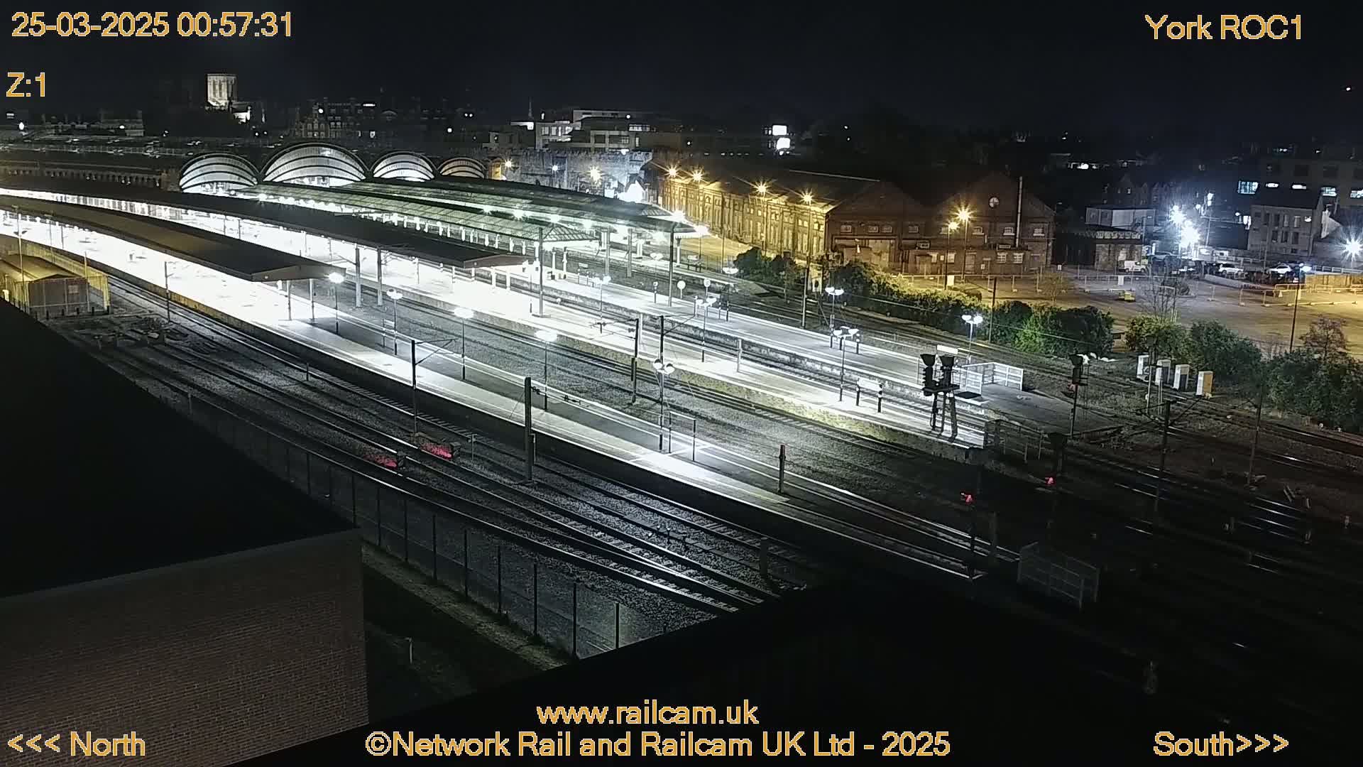 A nighttime, high-angle view of a brightly lit train station with numerous tracks and platforms.