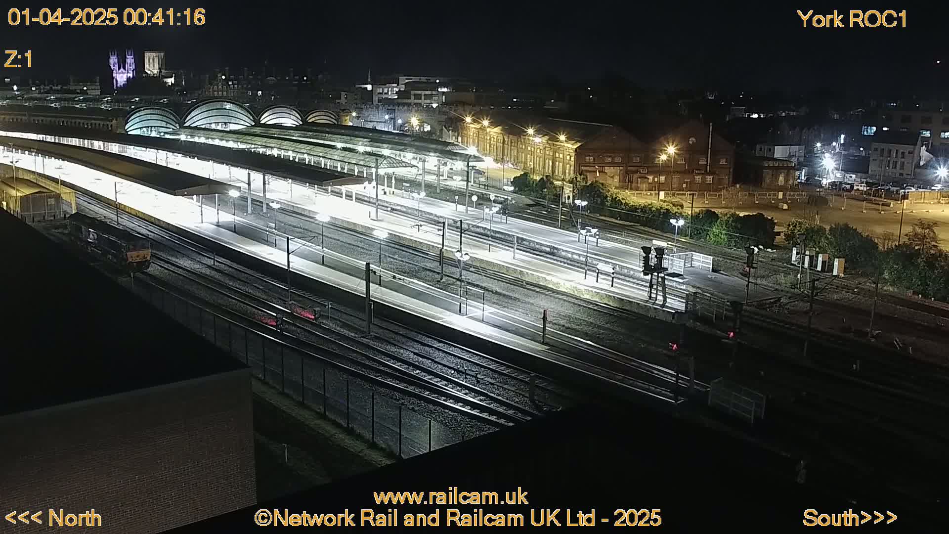 A brightly lit train station at night, with multiple train tracks and platforms, is viewed from above.