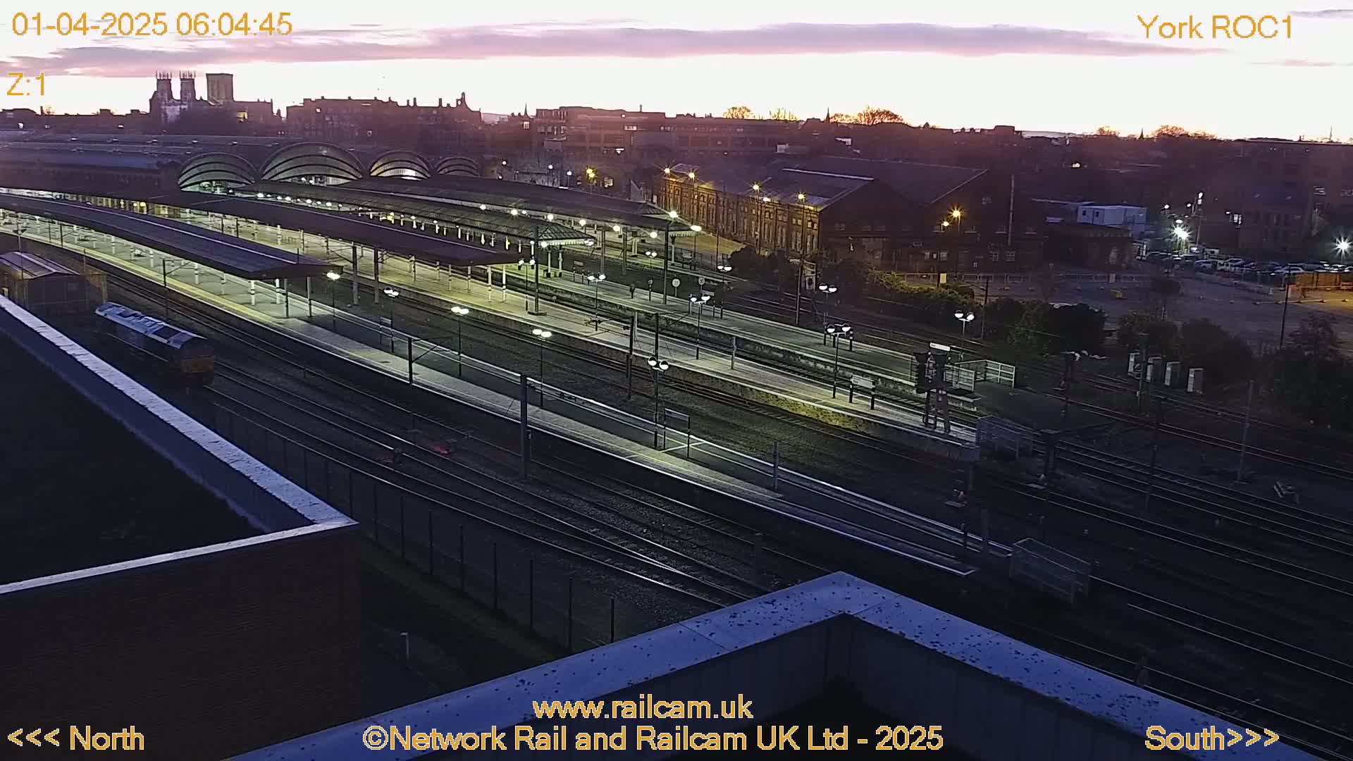 A high-angle, twilight view of a train station with multiple tracks and platforms, a locomotive, and surrounding city buildings under a dusky sky.