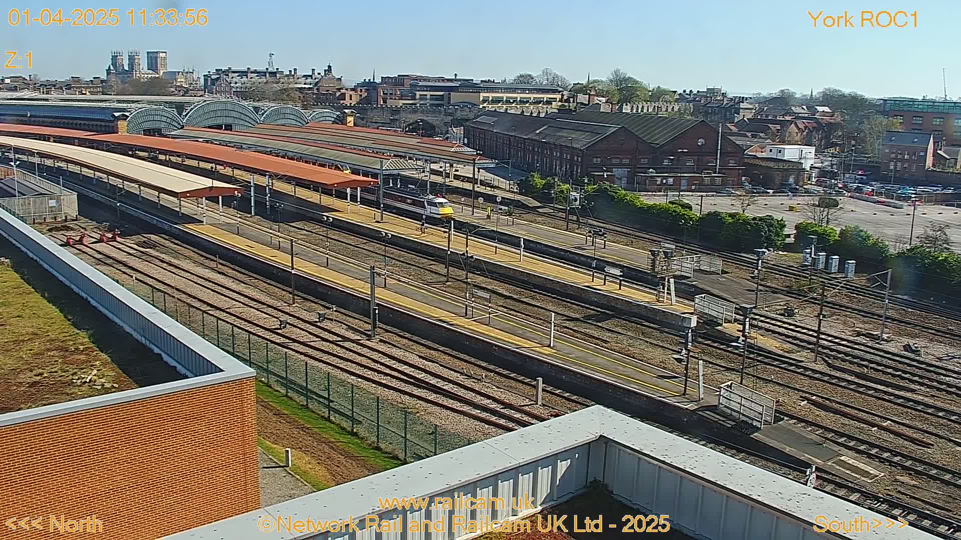 A high-angle, sunny view of a train station with multiple train tracks and platforms, a train visible on one track, and a cityscape in the background.
