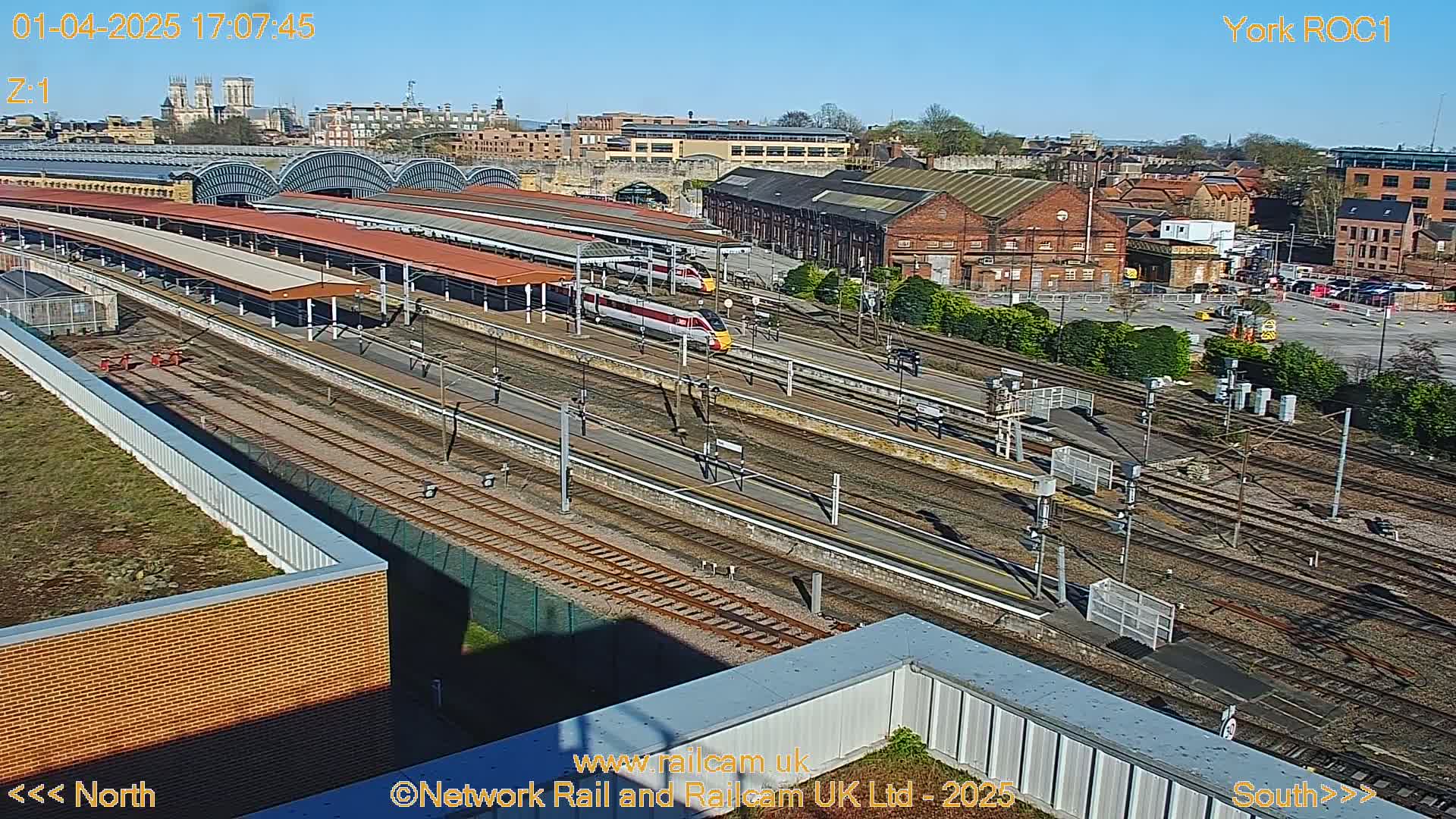 A high-angle view of a train station on a sunny day, showing multiple train tracks, platforms with covered waiting areas, and two high-speed trains.