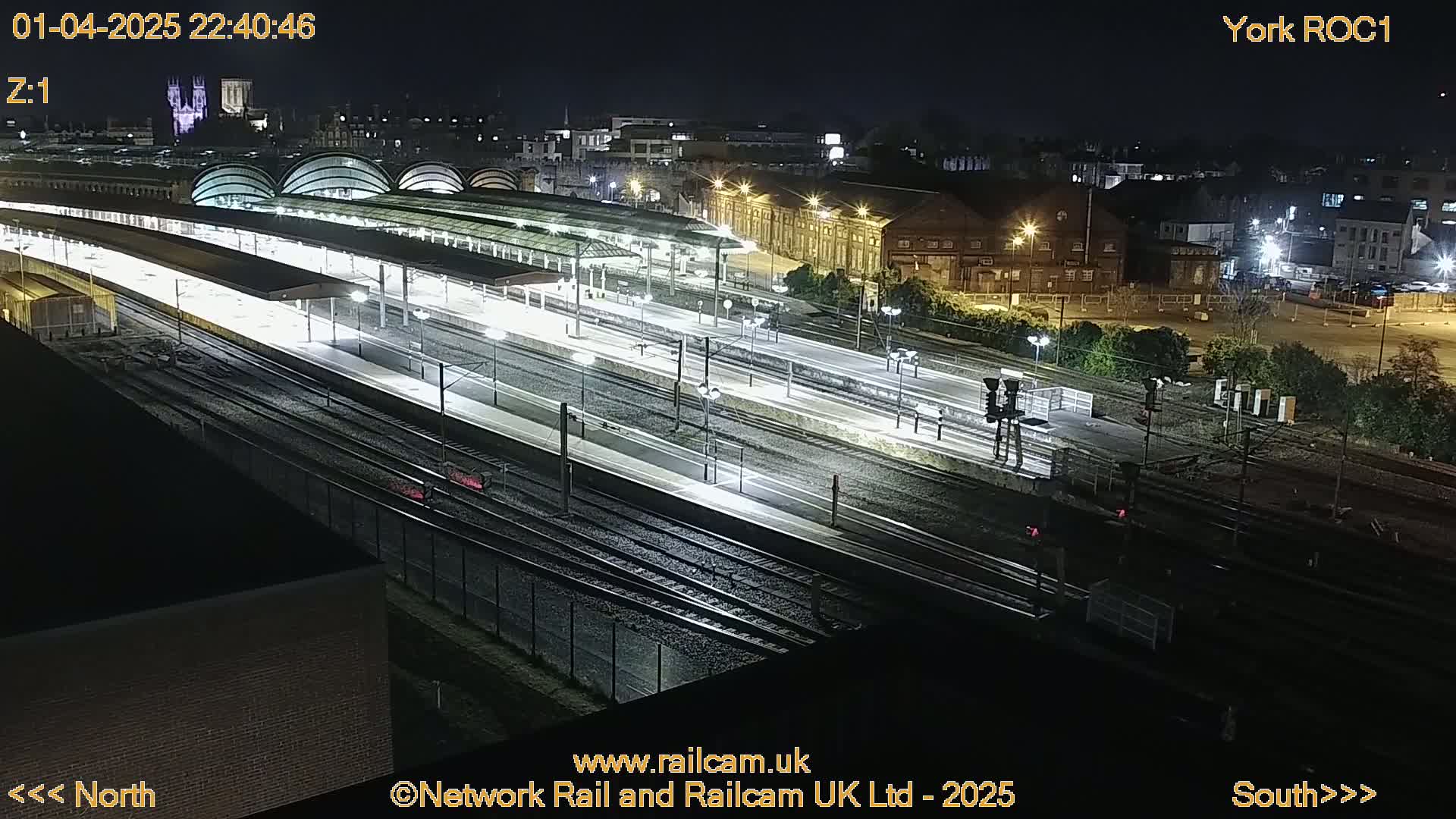 A nighttime, overhead view of a brightly lit train station with numerous tracks and platforms, surrounded by city buildings.