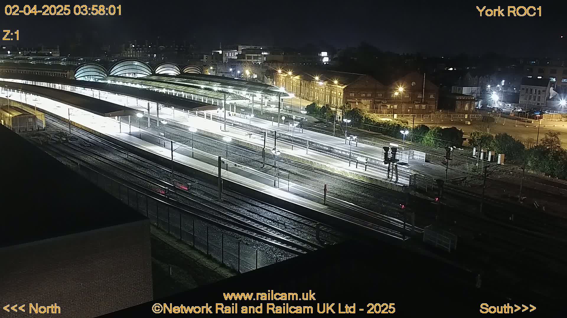A nighttime, high-angle view of a brightly lit train station with multiple tracks and platforms.