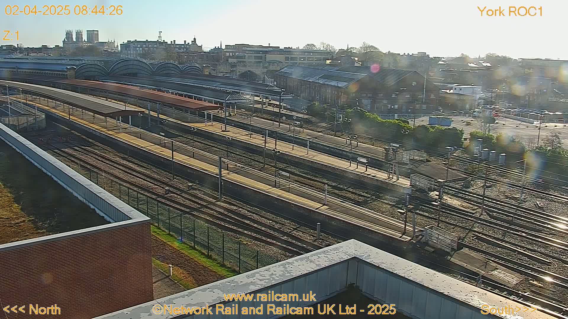 A high-angle, sunny view of a train station with numerous tracks and platforms, situated in a city with buildings visible in the background.