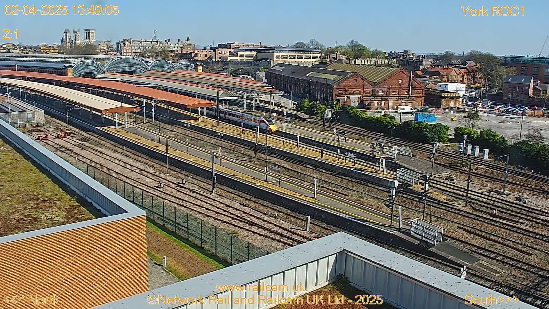 A high-angle view of a train station on a sunny day, showing multiple train tracks, platforms with covered waiting areas, and a high-speed train passing through.
