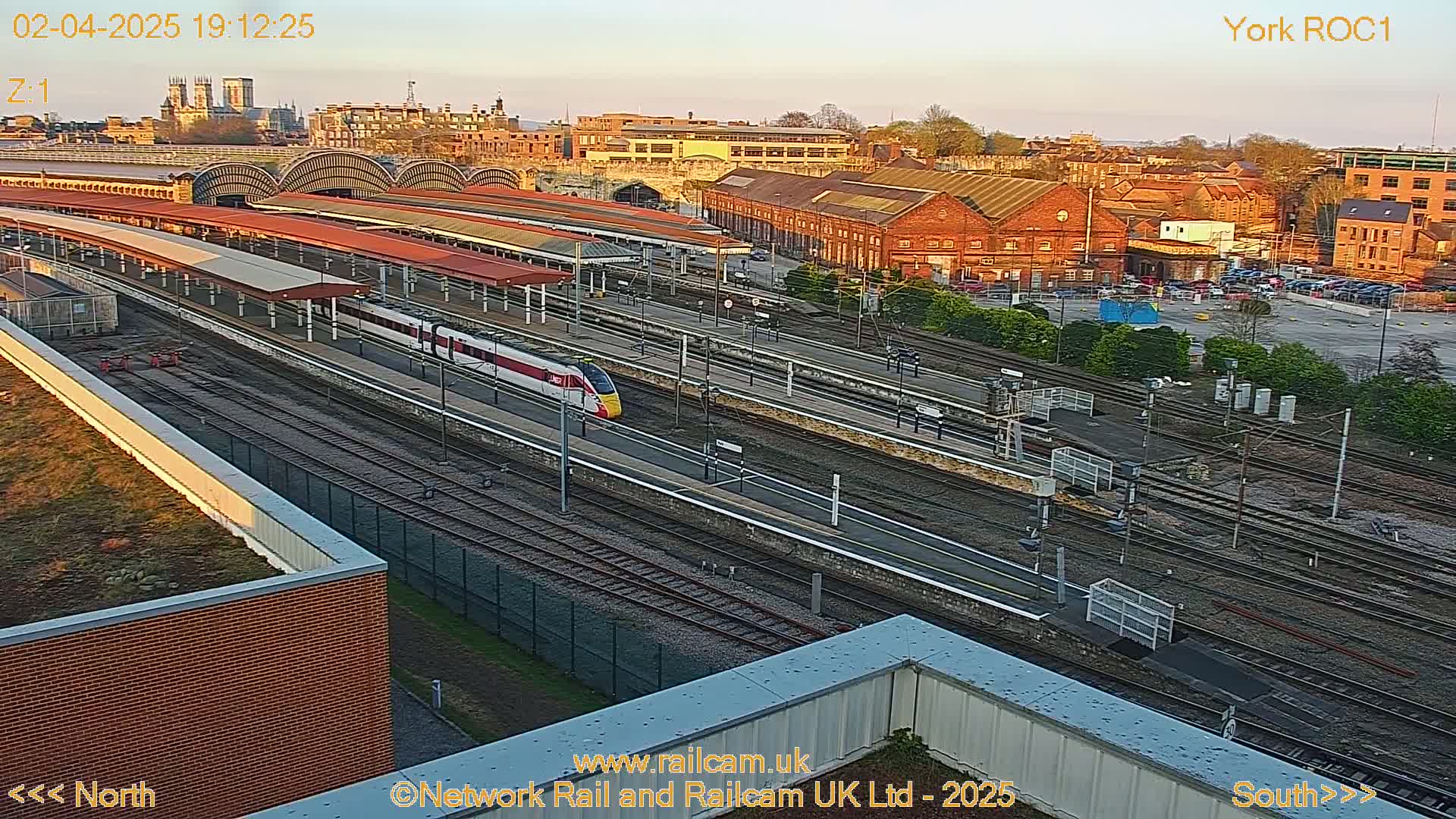 A high-angle, evening view of a train station with a high-speed train passing through numerous tracks, under a clear sky.