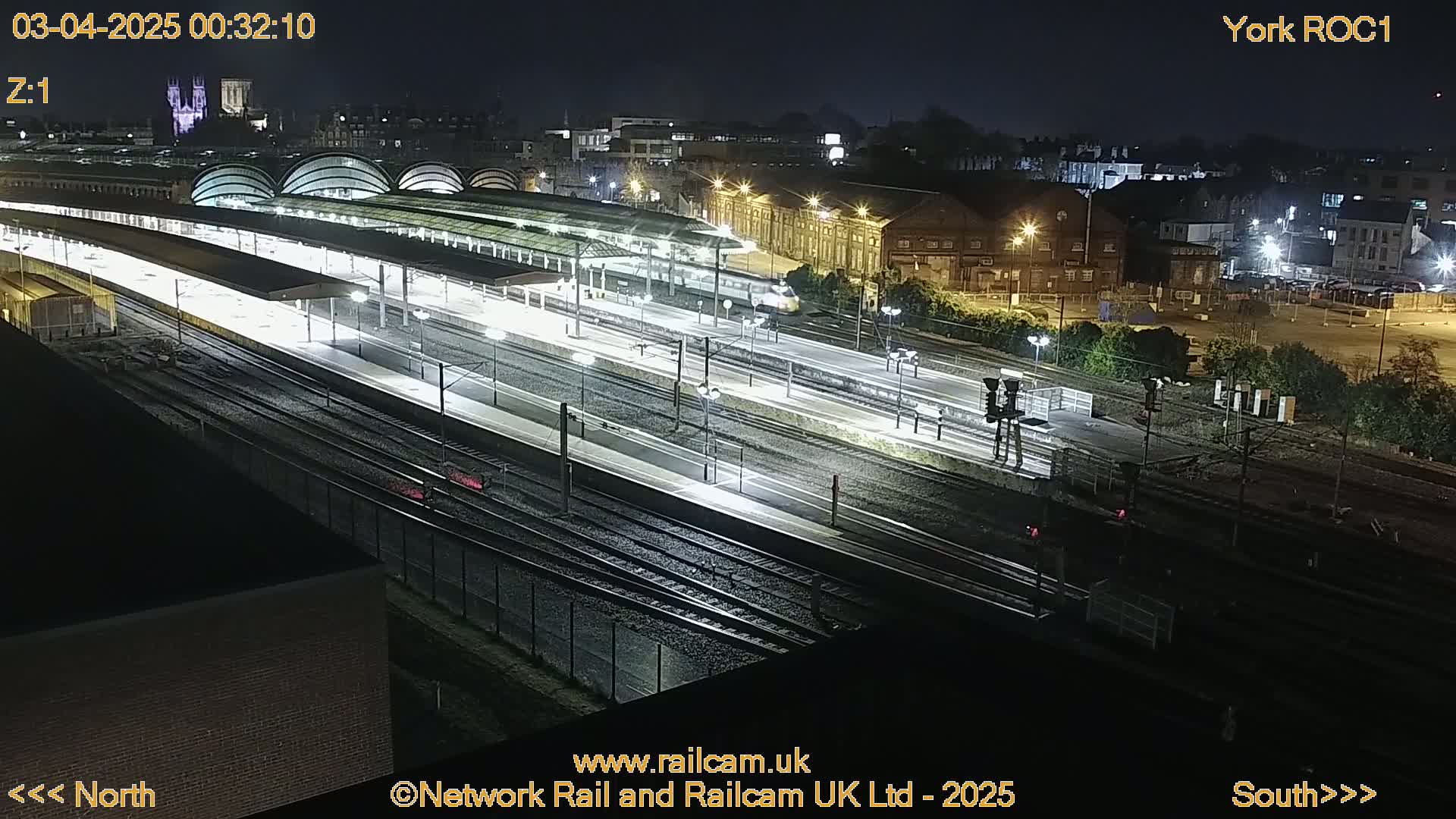 A nighttime, high-angle view of a brightly lit train station with multiple tracks and a train passing through.