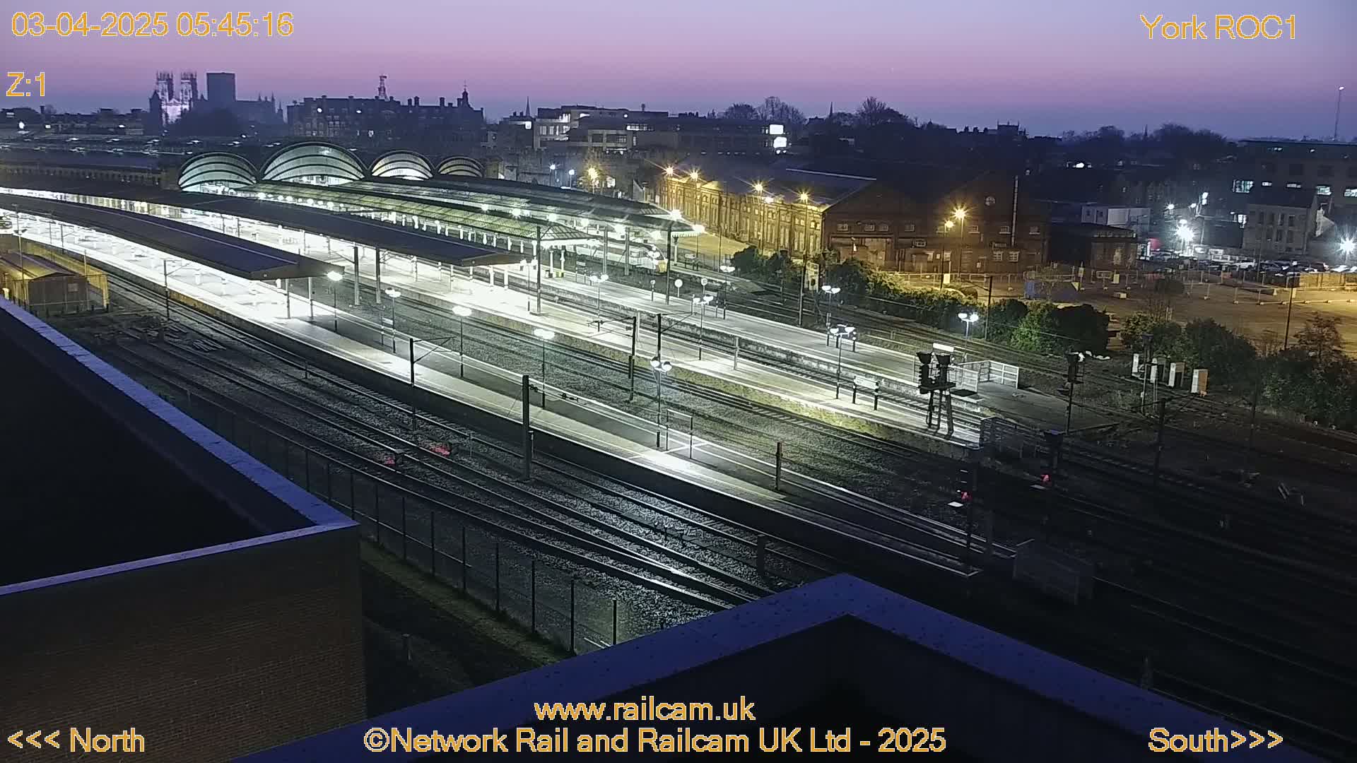 A nighttime, high-angle view of a train station with multiple tracks and platforms, illuminated by artificial lights under a twilight sky.