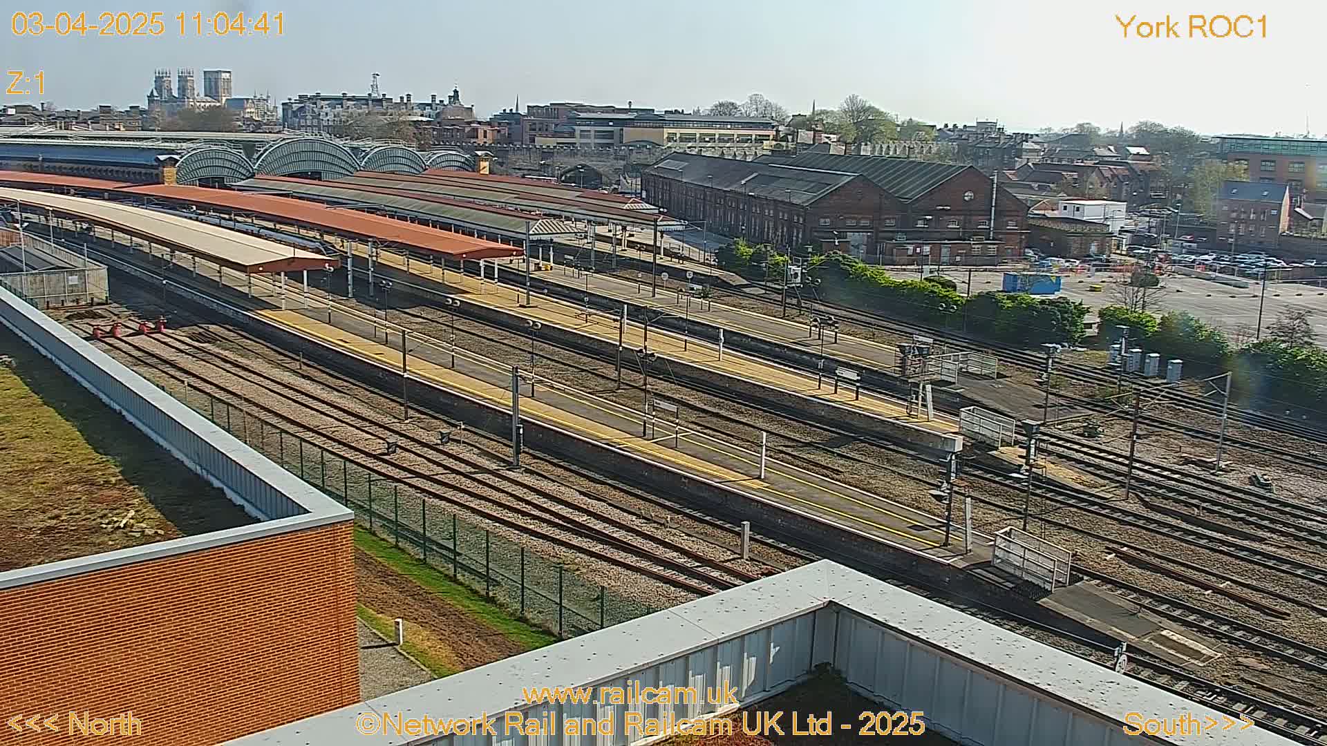 A high-angle view of a train station on a sunny day, showing multiple train platforms, tracks, and surrounding buildings.
