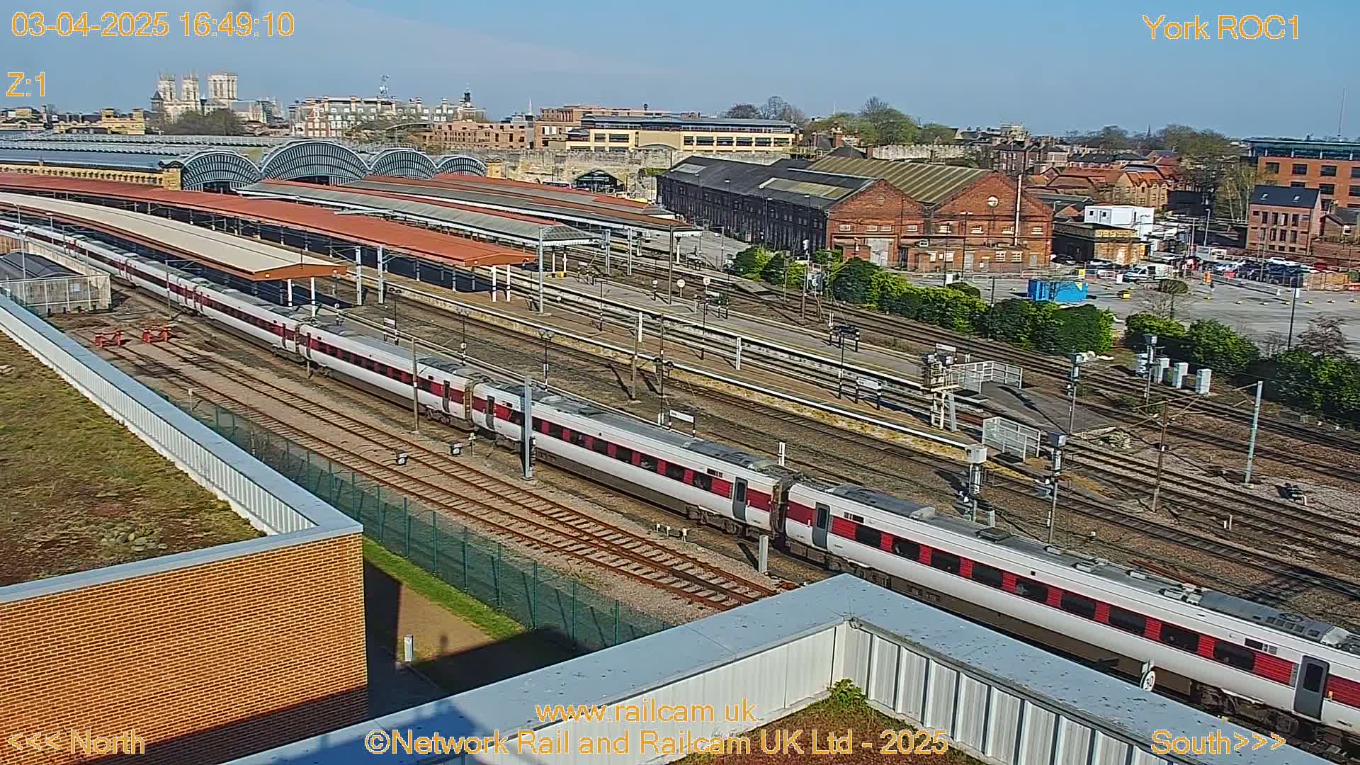 A long passenger train sits at a station on a sunny day, with a city skyline visible in the background.