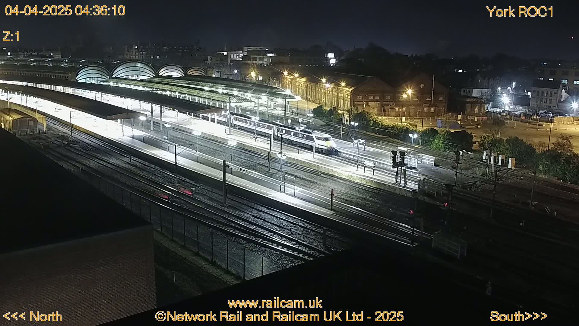 A high-angle nighttime view of a train station with a train departing, illuminated by artificial lights.
