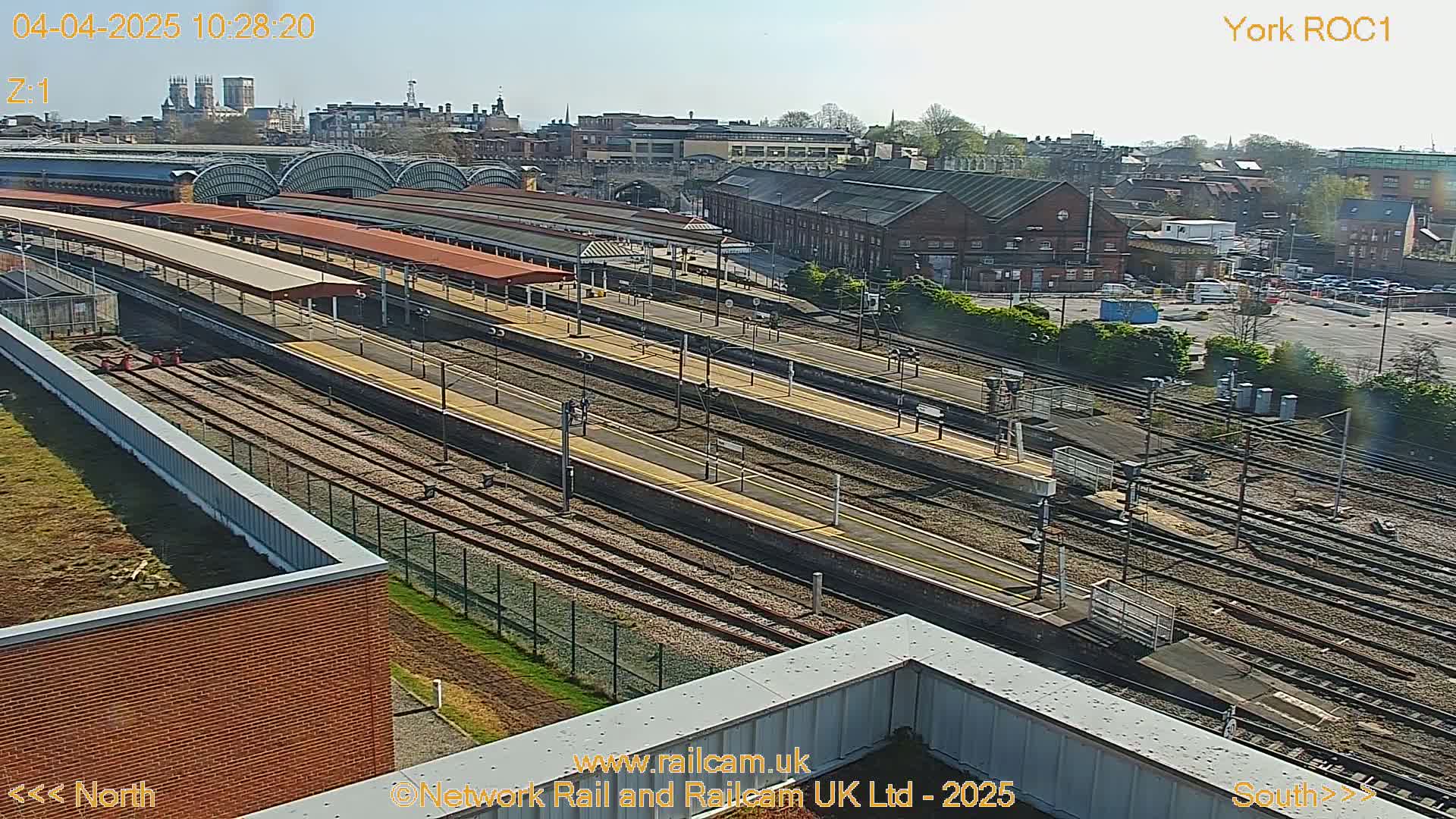 A high-angle view of a train station on a sunny day, showing multiple train tracks and platforms extending into the distance.