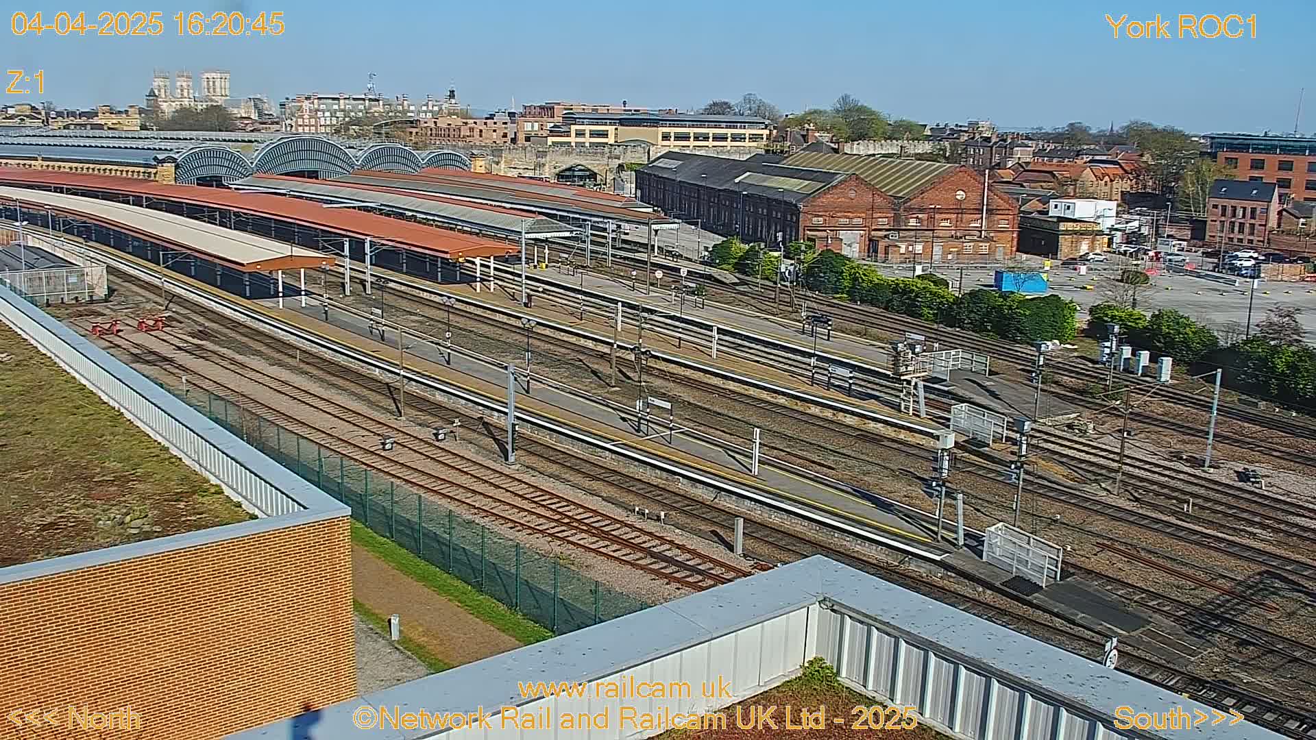 A high-angle, sunny view of a train station with numerous tracks, platforms, and surrounding buildings.