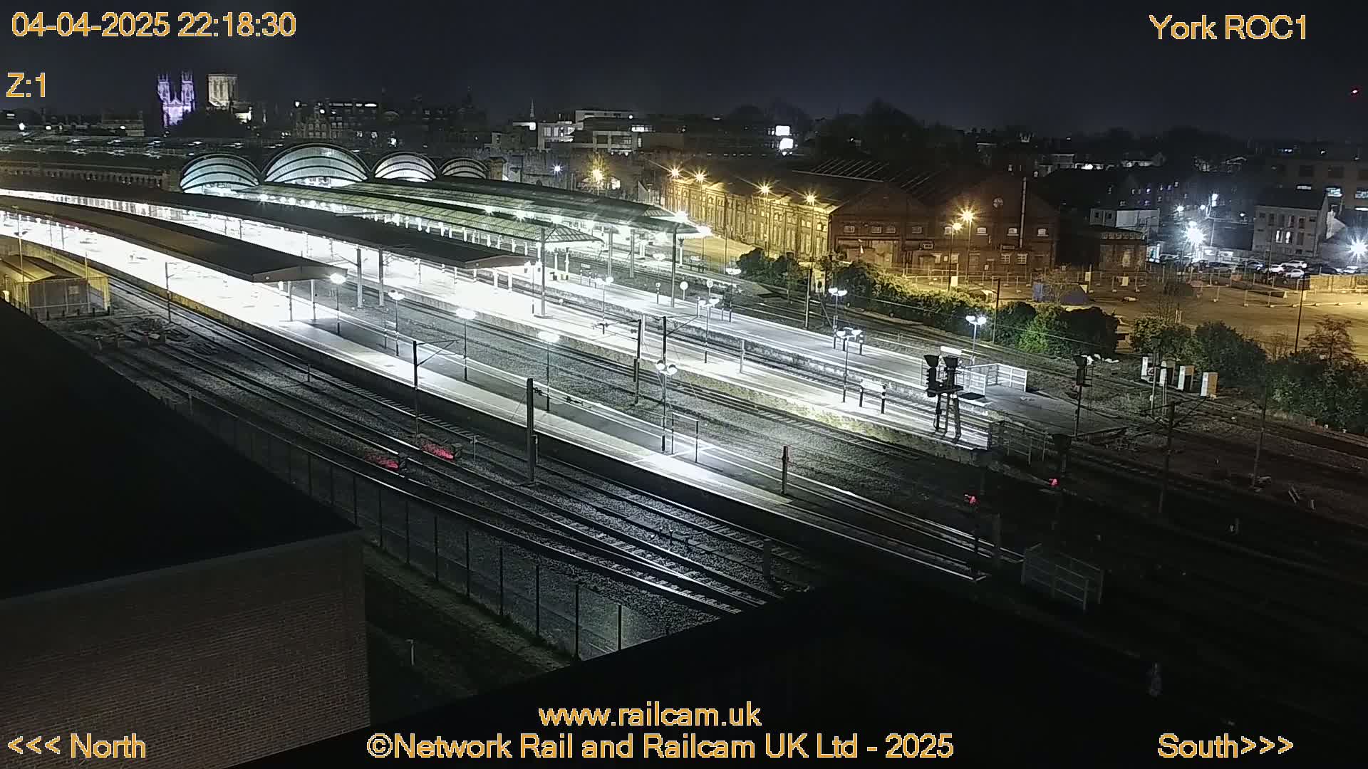 A nighttime, high-angle view of a train station with multiple tracks and platforms, illuminated by artificial lights.