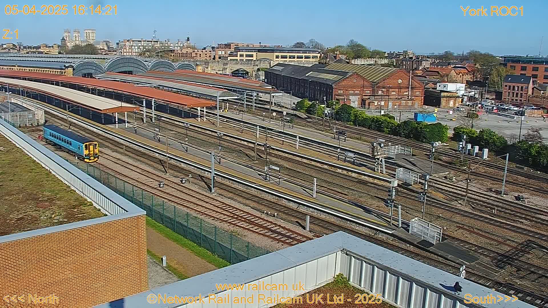 A blue train sits at a station platform amongst many railway tracks, under a sunny sky, with a city visible in the background.
