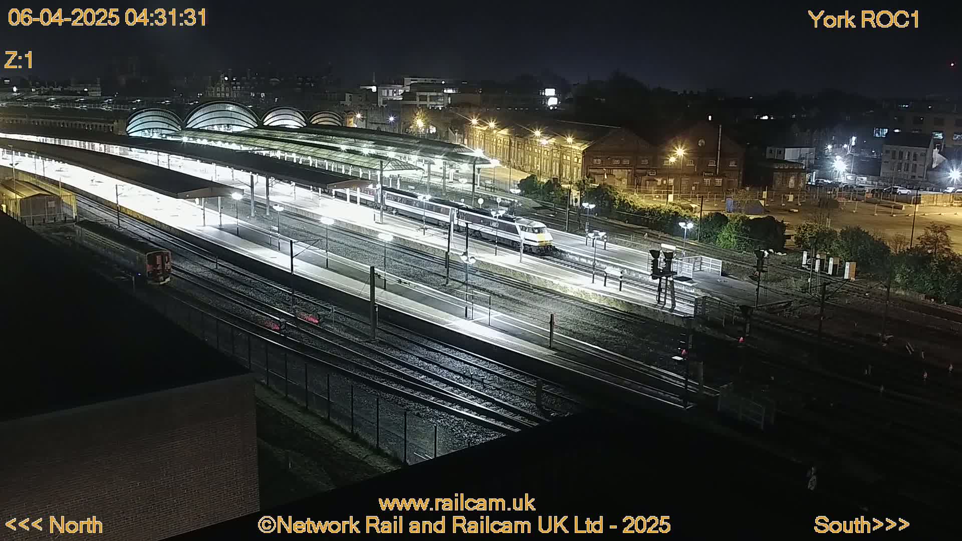 A high-angle night view shows a train passing through a brightly lit train station with multiple tracks and platforms.