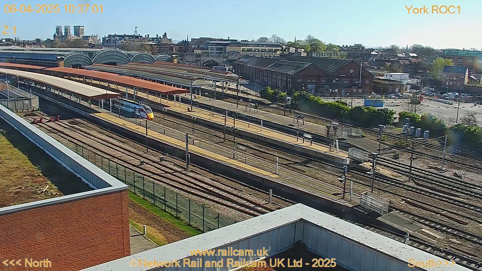 A high-angle, sunny view of a train station with a high-speed train arriving at a platform, surrounded by tracks and buildings.