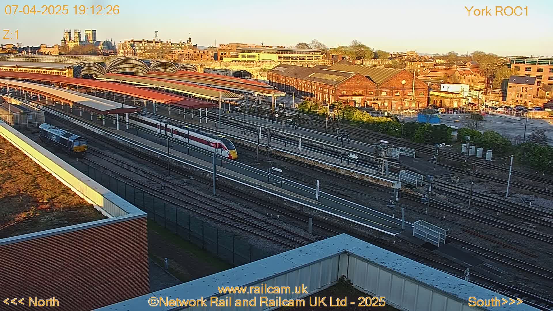 A high-angle, long shot of a train station with multiple train tracks and platforms, two trains, and surrounding buildings at sunset.