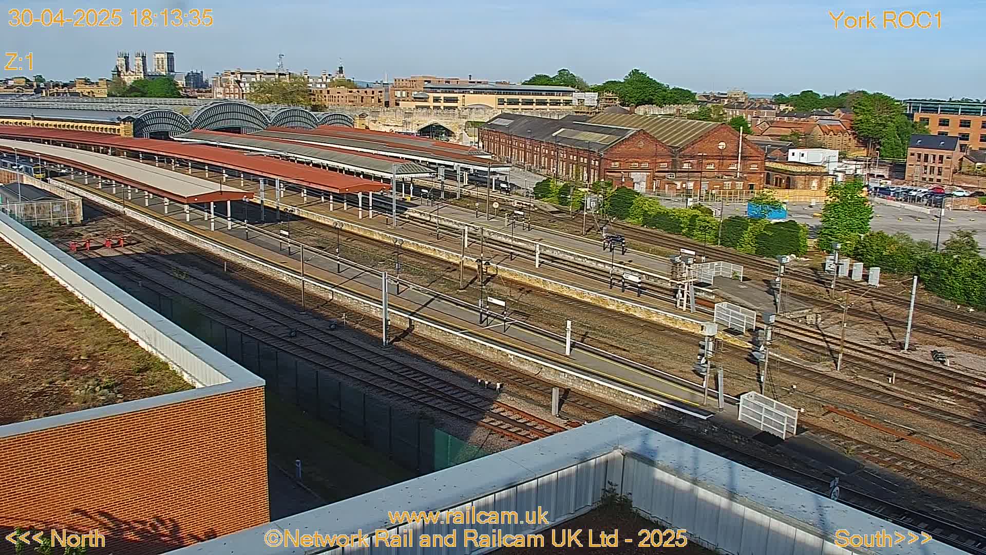 A high-angle, long shot of a train station on a sunny day, showing numerous train tracks and platforms, alongside adjacent buildings and a parking lot.