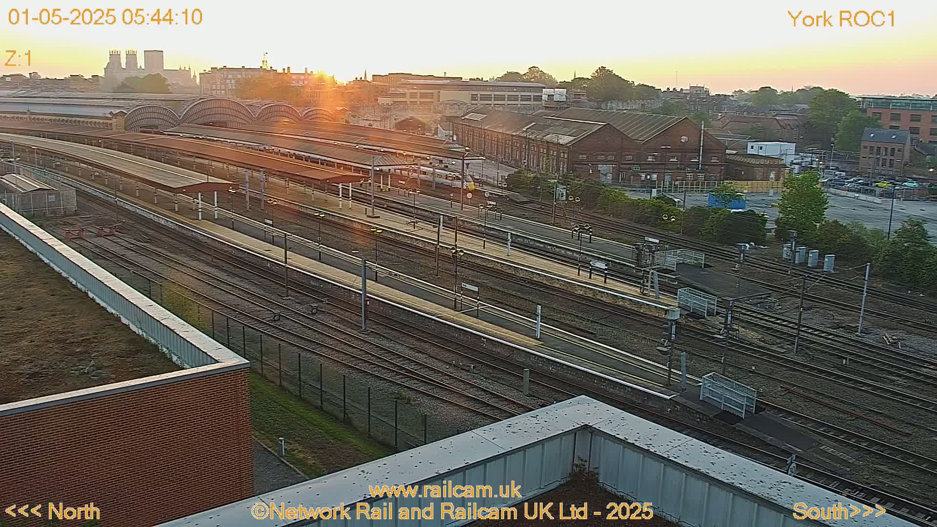 A sunrise illuminates a sprawling railway station with numerous tracks and platforms, surrounded by industrial buildings and a parking lot.