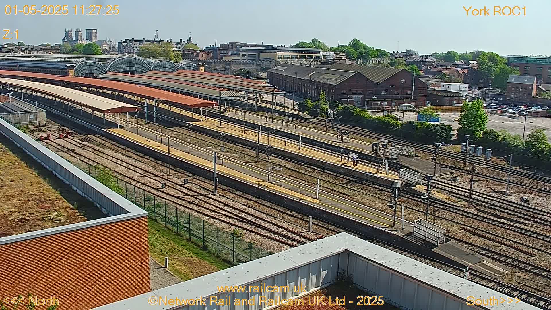 A high-angle, sunny view of a railway station with multiple tracks, platforms, and surrounding buildings.