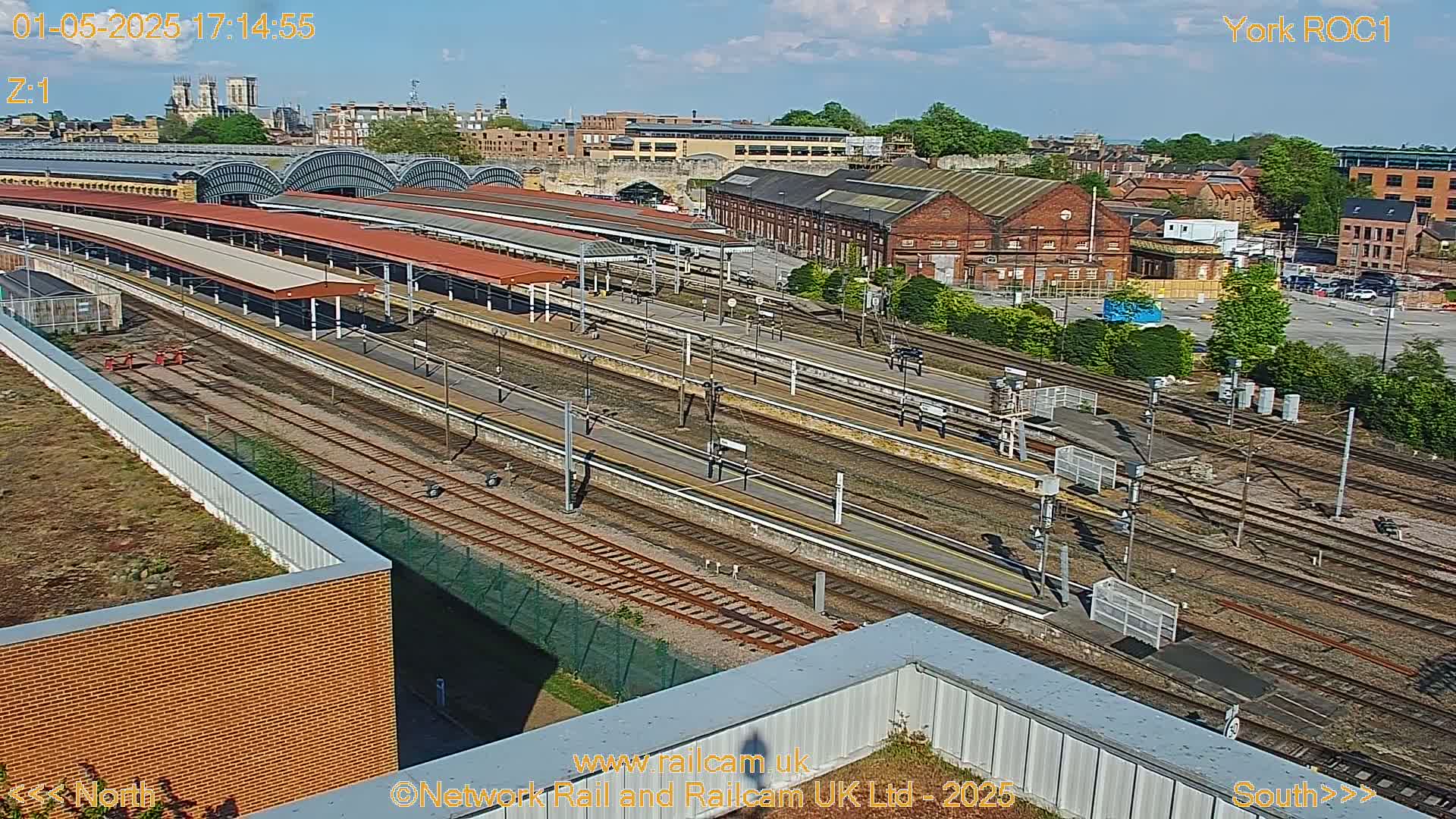 A high-angle, sunny view of a train station with multiple tracks and platforms, surrounded by industrial buildings and greenery.