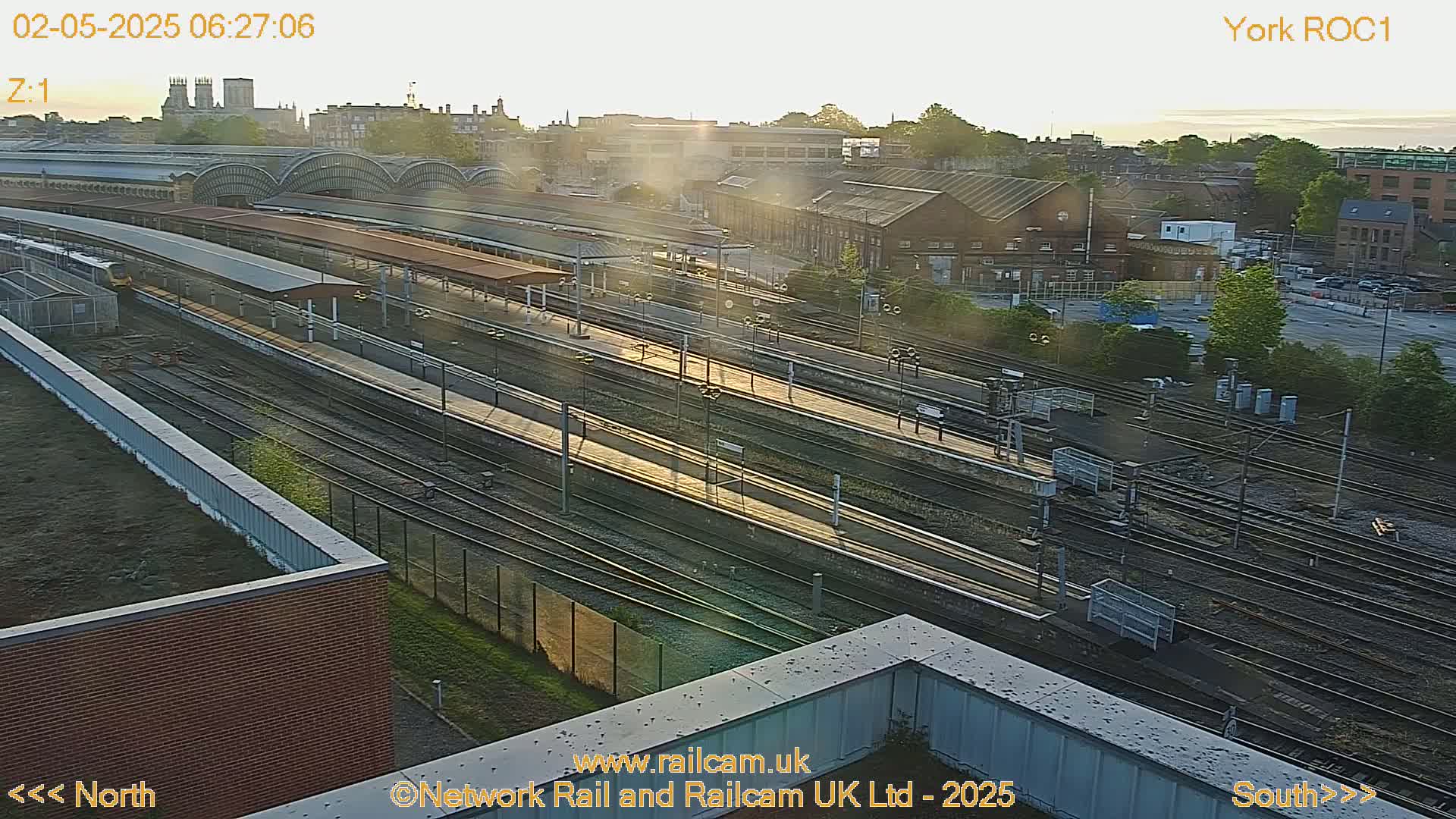 A high-angle, long shot of a train station at sunrise, showing numerous train tracks and platforms with a few trains visible, alongside surrounding buildings.