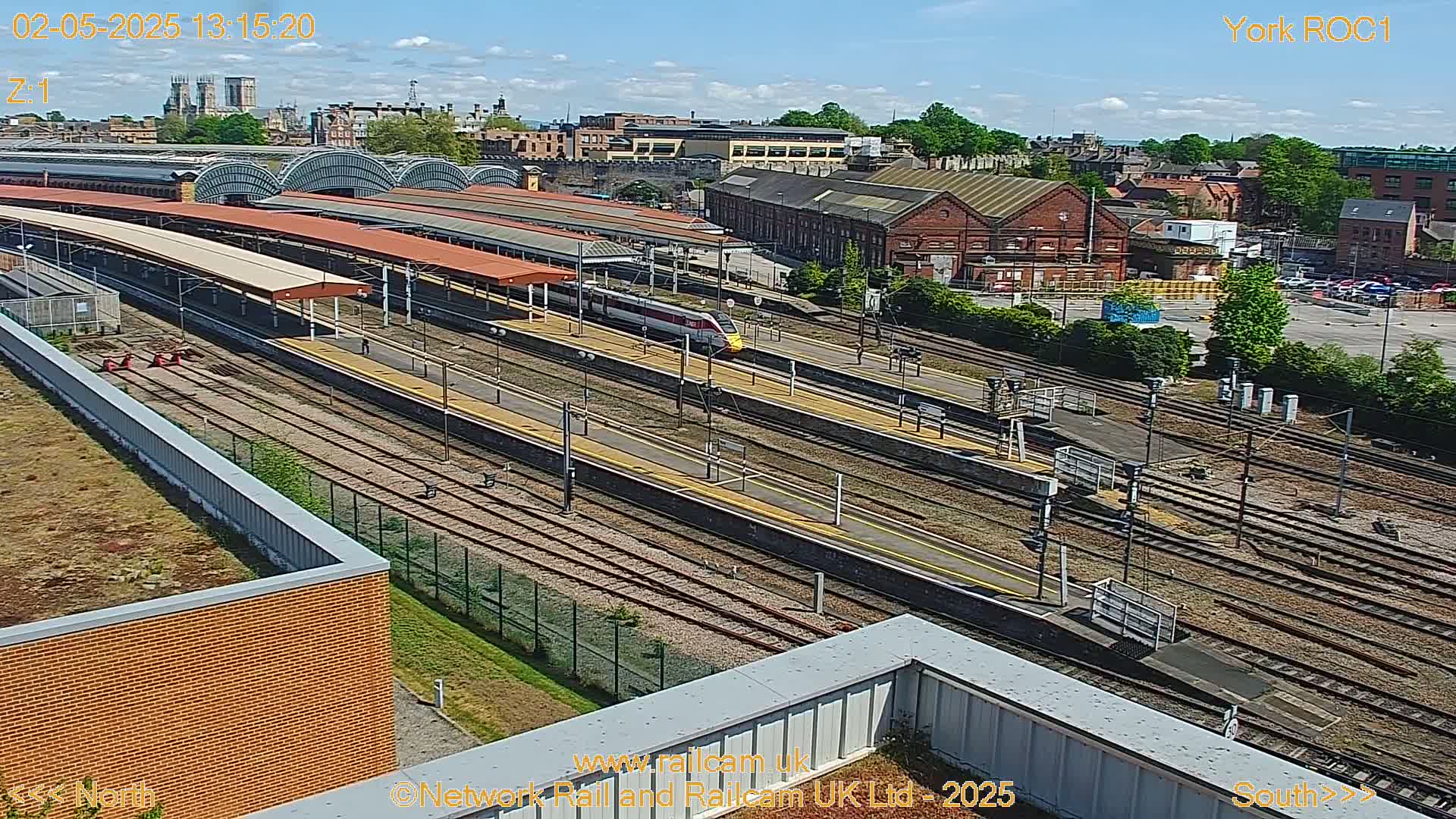 A high-angle, sunny view of a train station with a high-speed train passing through numerous train tracks and platforms, surrounded by industrial buildings and greenery.