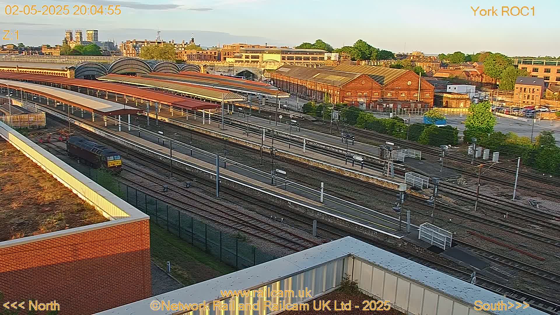 A high-angle, long shot of a train station at dusk, showing multiple train tracks, platforms, and surrounding buildings, with a locomotive visible on one of the tracks.