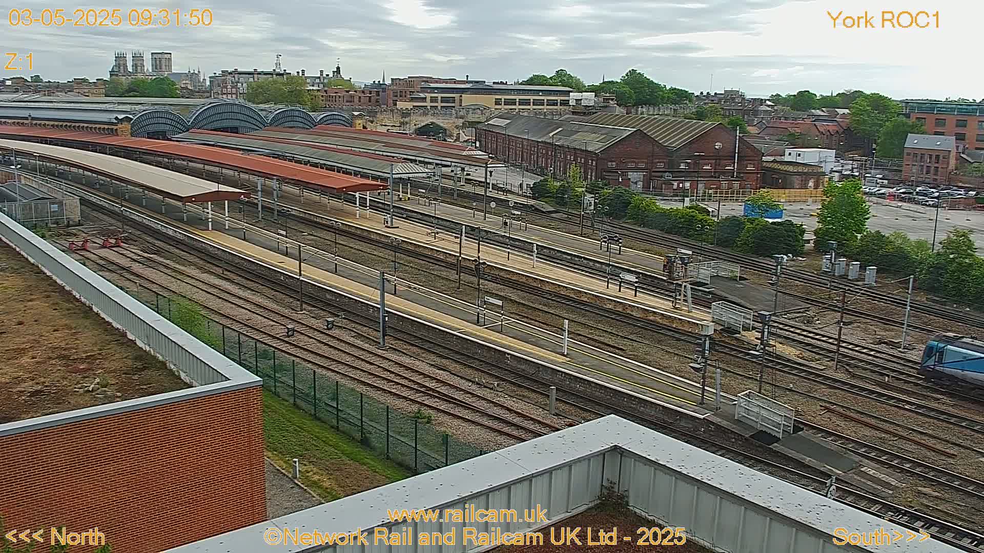 A high-angle, long shot of a railway station with multiple train tracks and platforms under an overcast sky.