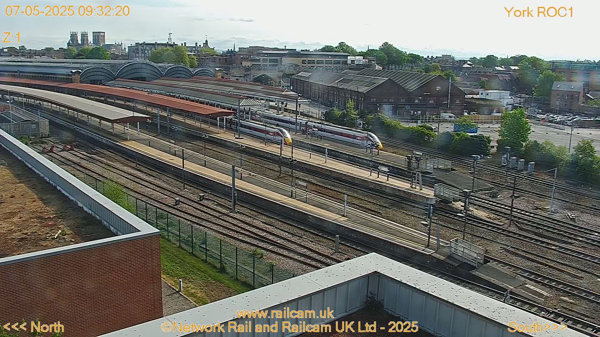 A high-angle, long shot of a train station on a sunny day, showing two high-speed trains at the platform and numerous railway tracks.