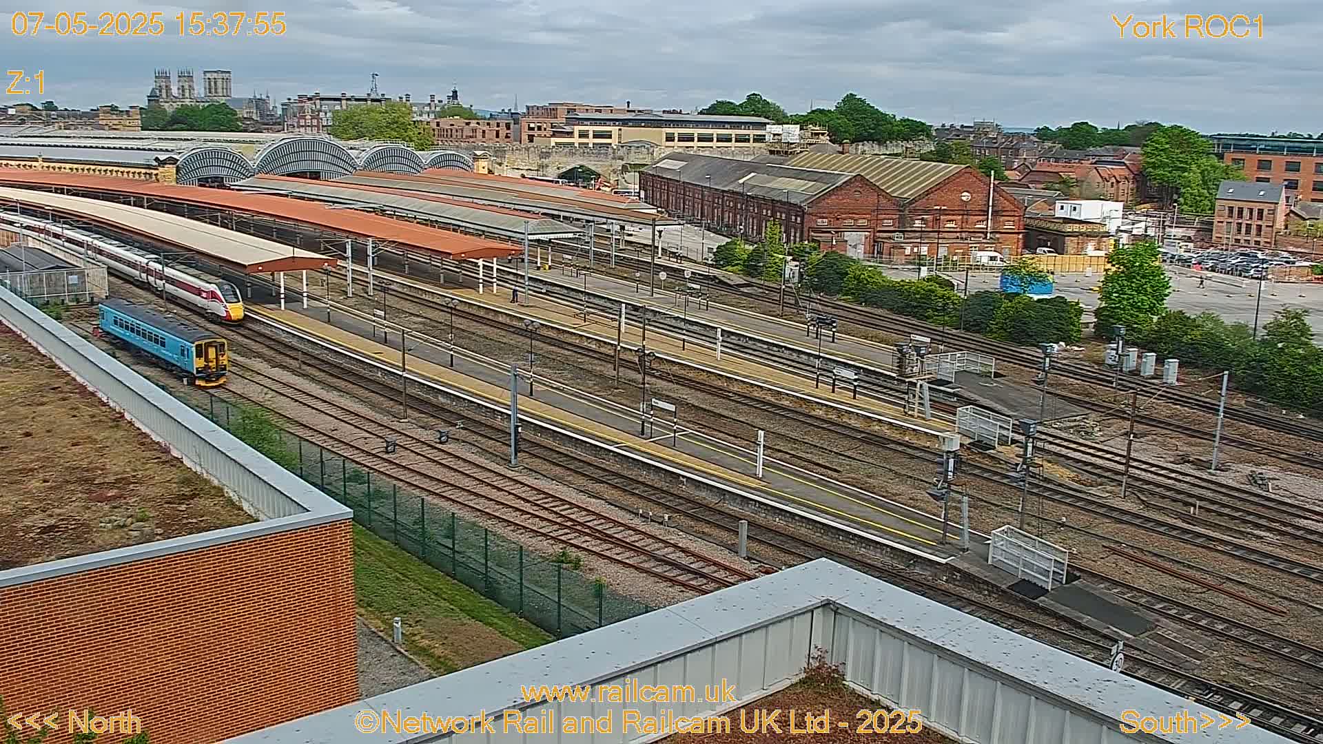 A high-angle view of a train station on an overcast day shows several trains and tracks, with a city in the background.