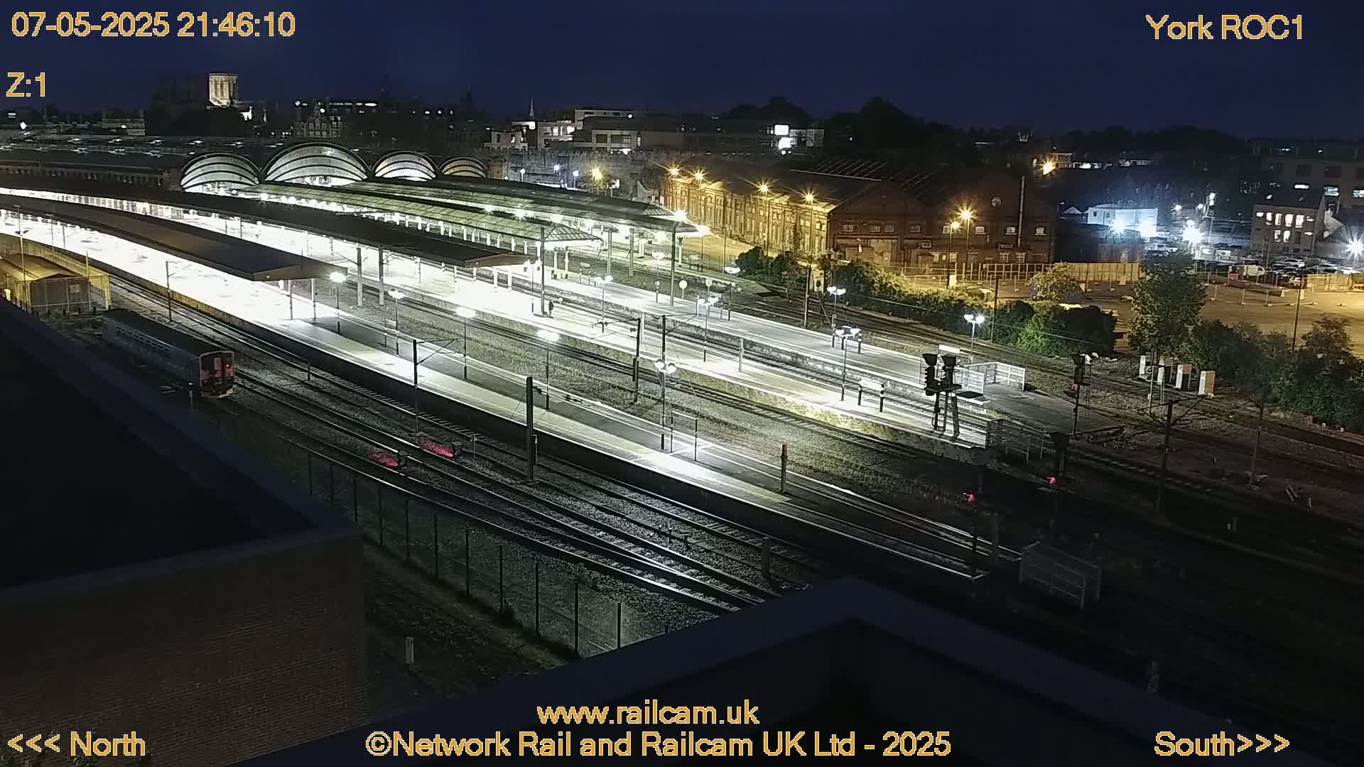 A nighttime, high-angle view of a well-lit train station with multiple tracks and platforms.
