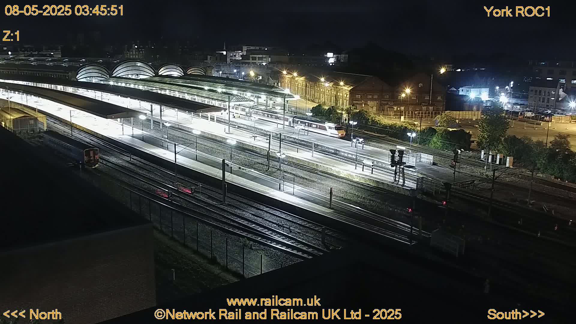A brightly lit train station at night, with a train on the tracks and several platforms visible under curved roofs.